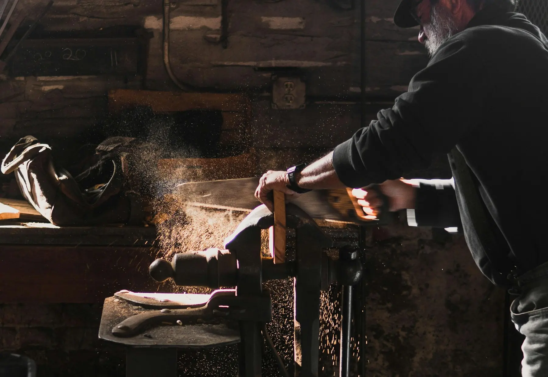 Ein Schreiner sägt im Gegenlicht ein Werkstück im Schraubstock, der Sägestaub leuchtet.